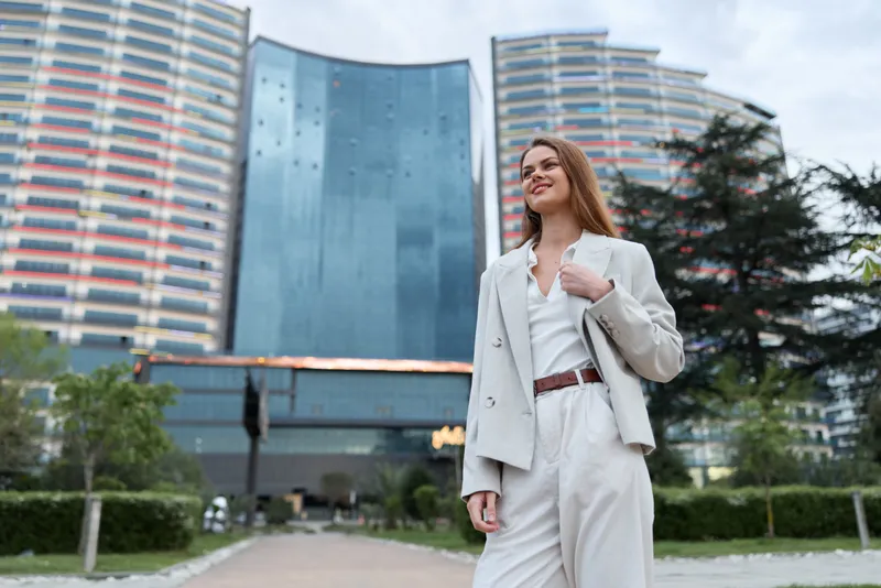 confident woman in beige business suit standing outside modern office building with skyscrapers and greenery