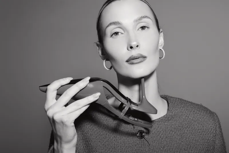 Elegant white woman with fair skin holding black high heel shoe near face in studio. Fashion and style concept.