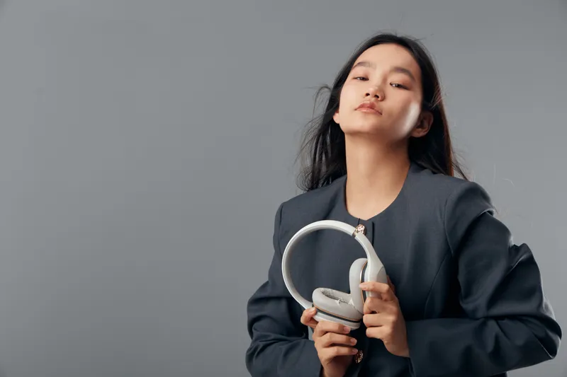 Young woman holding white headphones with closed eyes in studio, wearing dark oversized jacket on gray background, posing with confident and relaxed expression for music or technology concept.