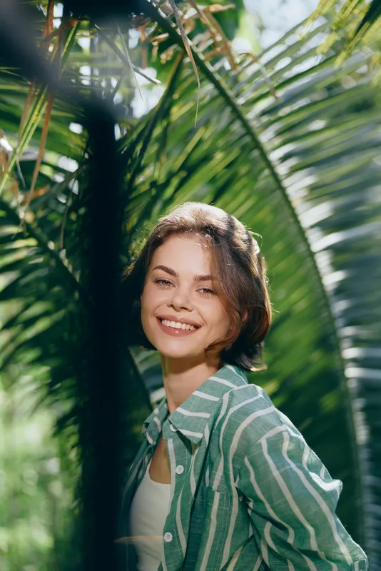 Smiling young woman posing amid lush tropical foliage and palm leaves, bathed in soft sunlight, wearing a striped green shirt.