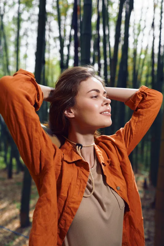 Woman forest portrait relaxing with hands behind head and smile, wearing orange jacket in sunlit woodland. Outdoors lifestyle, nature escape, peaceful young adult enjoying fresh air.