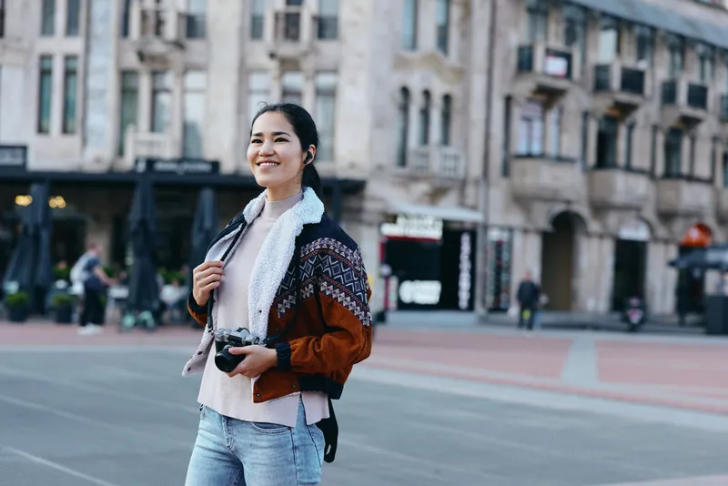 Woman photographer on a street, camera in hand, in urban outdoor setting, smiling and stylish in a casual jacket, capturing city life and travel moments