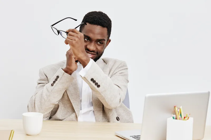 Professional portrait of a smiling young man with dark skin, wearing a light beige blazer, holding eyeglasses, sitting at a desk with a white laptop and a coffee cup against a plain white background. 