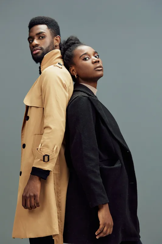 Stylish couple standing back to back in modern fashion coats, showcasing confident expressions against a gray background for a trendy photoshoot