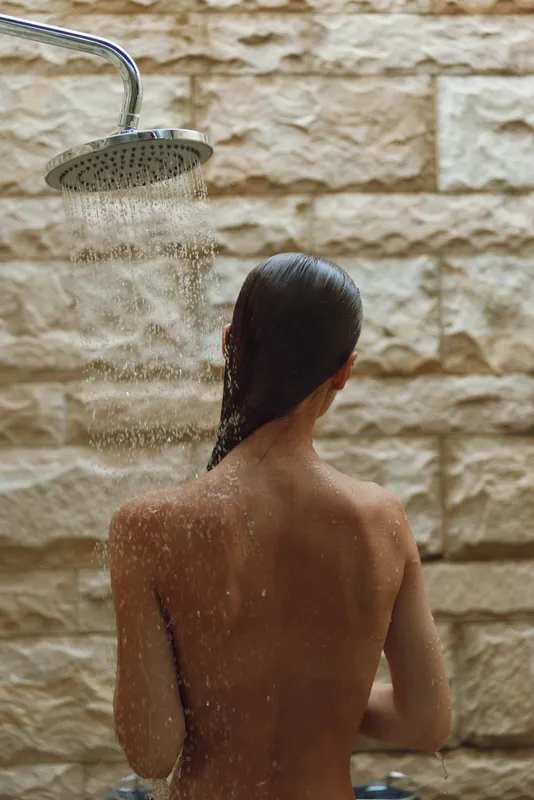 woman showering under rainfall showerhead with wet hair against a textured stone wall, wellness, relaxation, indoor bathing, natural light