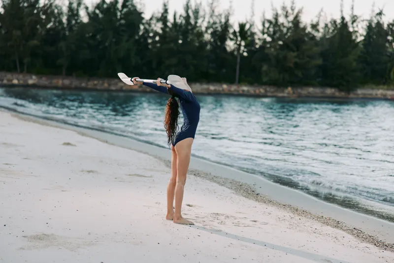 A girl on a sandy beach stretches arms overhead, wearing a dark swimsuit and wide brim hat, facing calm water under soft natural light.
