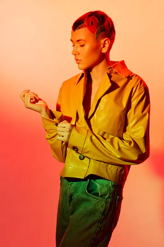 young woman with short styled hair wearing yellow jacket and green pants posing thoughtfully against warm gradient background with dramatic lighting