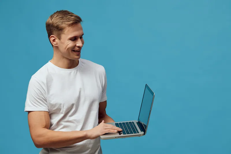 Smiling young caucasian man working with laptop wearing white t-shirt looking at screen on blue background digital nomad concept technology communication