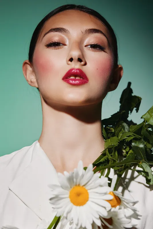 Young woman with glossy red lips and shiny eyeshadow poses confidently with vibrant greenery and daisies. Creative beauty portrait with fresh fashion vibes on a green background.