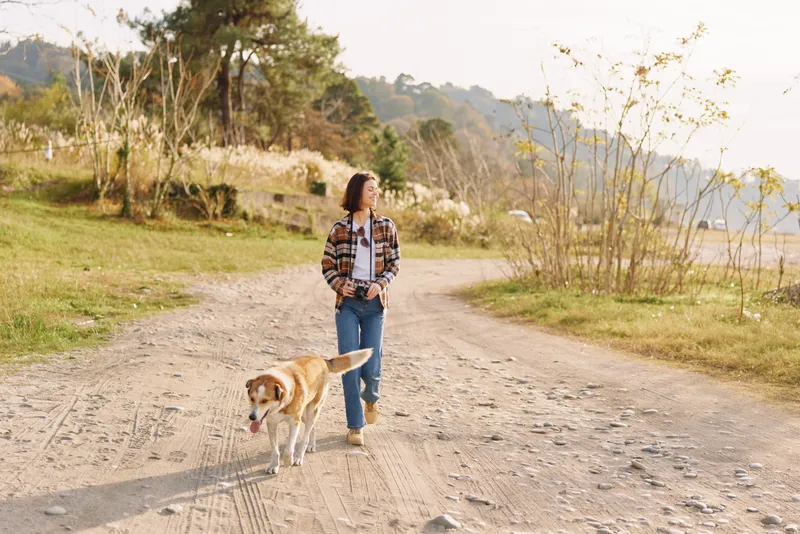 Woman walking along a dirt path with a dog in a sunlit park, casually dressed in jeans and a plaid shirt. Wide scenic trail, trees in bloom, peaceful outdoor moment.
