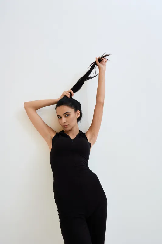 Young woman in black sleeveless outfit poses confidently against white background, holding her long dark hair with one arm raised, showcasing elegance and strength.