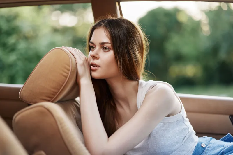 Young woman lost in thought, resting on car seat, wearing a casual white tank top, with a serene expression in nature background, summer vibes, car travel.