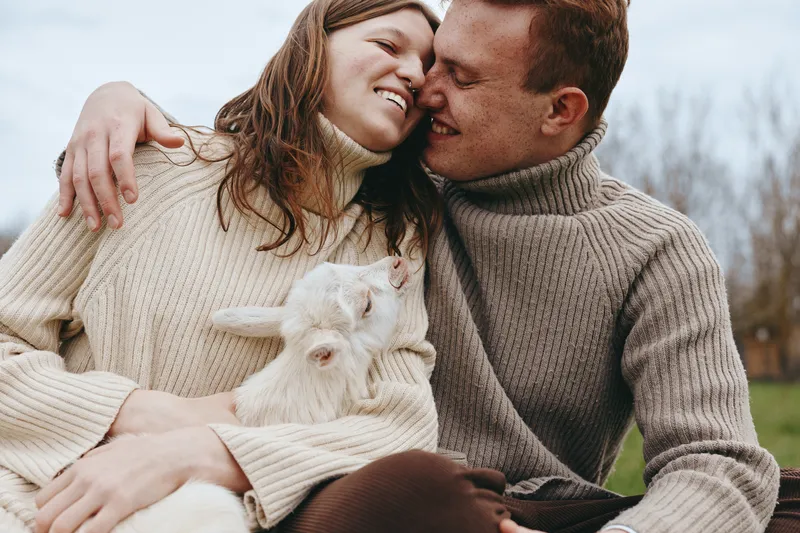 Couple in cozy sweaters cuddle a white goat kid outdoors, smiling and affectionate. Natural light highlights warmth, connection, and a simple lifestyle moment in the countryside.