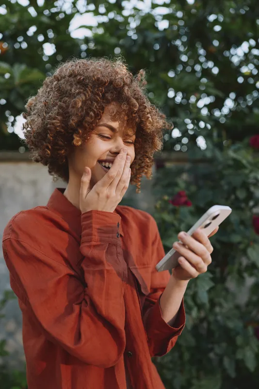 Cheerful woman lifestyle shot outdoors, smiling while checking her smartphone, wearing a rust sweater, lush garden backdrop, candid moment of digital connection