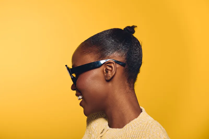 Profile of a smiling black woman with dark skin wearing black sunglasses and a beige sweater against a yellow background. Concept of fashion, confidence, and lifestyle portrait.