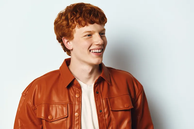 Young man with red hair smiling looking sideways in brown leather jacket and white shirt against plain white background in studio portrait shot.