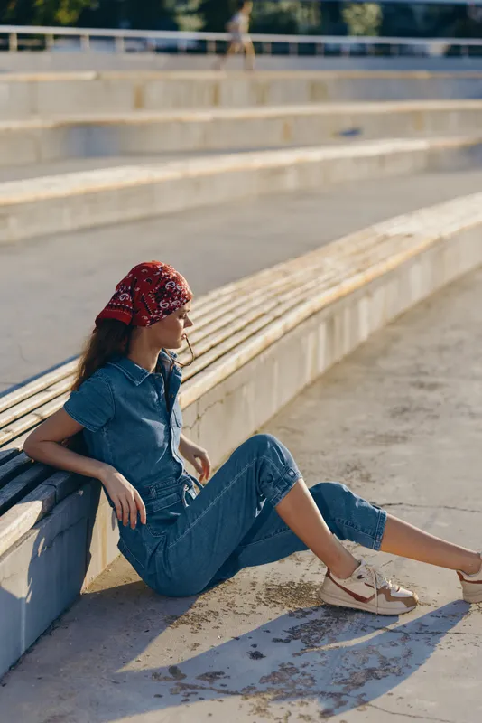 young woman wearing denim outfit and red headscarf sitting outdoors on sunny day near stone steps in casual style