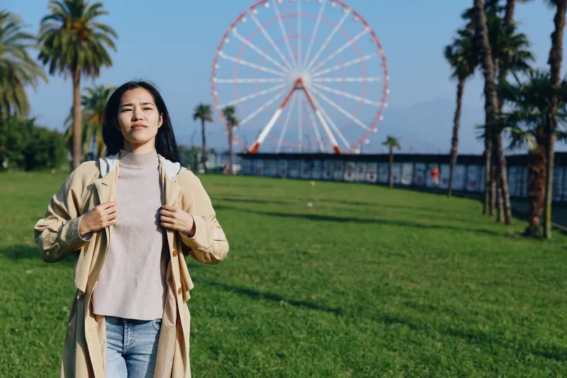 Woman in beige coat and knit sweater stands in a sunny park with a large ferris wheel in the background, palm trees frame the scene, a relaxed moment of outdoor leisure and city views