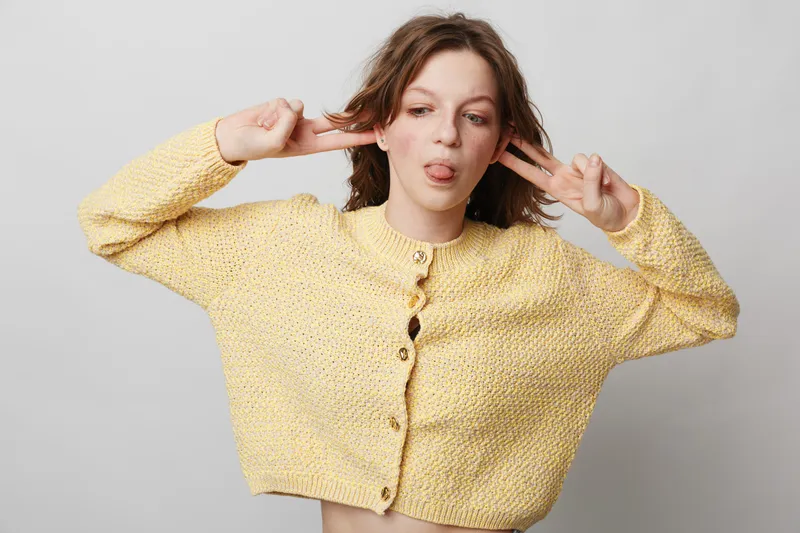 Young woman in yellow knitted sweater makes playful face with tongue out and fingers near ears. Studio portrait on grey background. Concept of fun, youth, casual style, and carefree mood.
