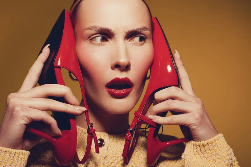 Woman with red lipstick holds red high heels near face, wearing yellow sweater. Concept of fashion, style, beauty, and bold accessories on warm background.