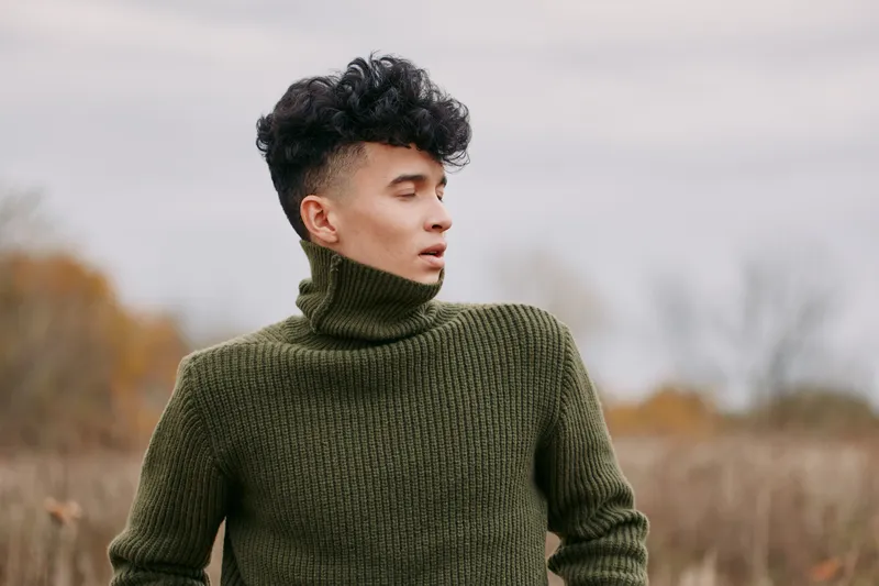 A candid outdoor portrait of a young man in a moss green turtleneck, looking thoughtfully to the side, highlighting authenticity and genuine emotion with tactile knit detail and natural expression.