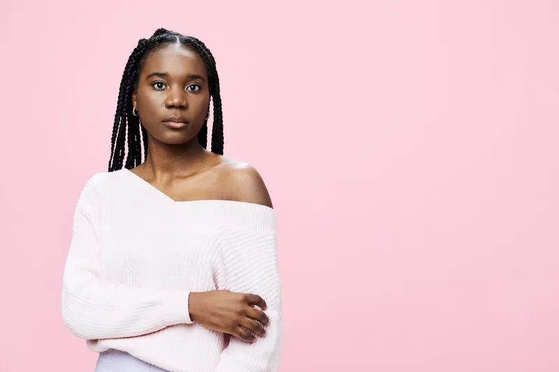 Young Black woman in a soft pink sweater with casual style, exhibiting a serious expression against a pastel pink background, ideal for lifestyle and fashion concepts