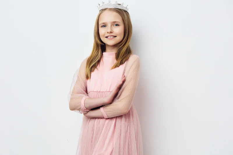 A cheerful young girl in a pink dress with tulle sleeves, wearing a tiara and smiling confidently against a simple white background