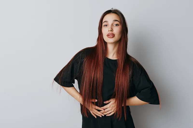Young woman with long hair and casual black t shirt standing with hands on waist against white background. Female model posing with serious expression and natural makeup in studio.