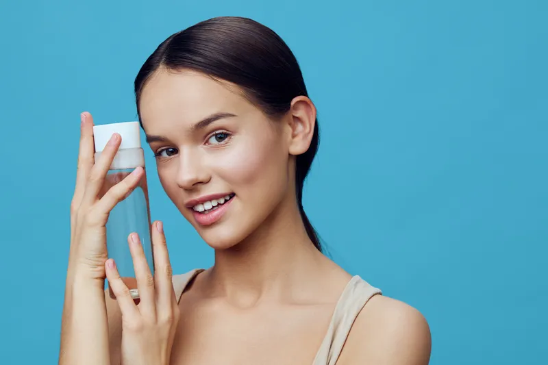 Young woman with clear skin holding a skincare bottle against a blue background, showcasing confidence and beauty in a minimalist style
