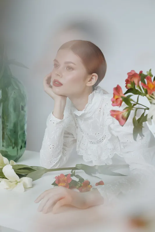 Elegant young woman with short hair in a white blouse, surrounded by flowers and soft colors, reflecting tranquility and beauty in a serene setting