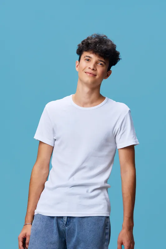 young man wearing white t-shirt and blue jeans smiles gently standing against solid light blue background with curly hair and relaxed posture