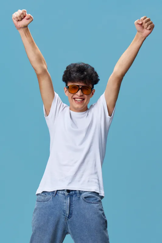 Happy young man raising arms in celebration wearing sunglasses and casual outfit against blue background, expressing joy and excitement.