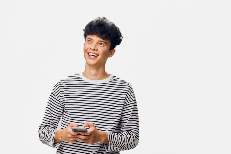 young man smiling joyfully while holding smartphone, wearing casual striped shirt, looking away on white background, happy communication concept