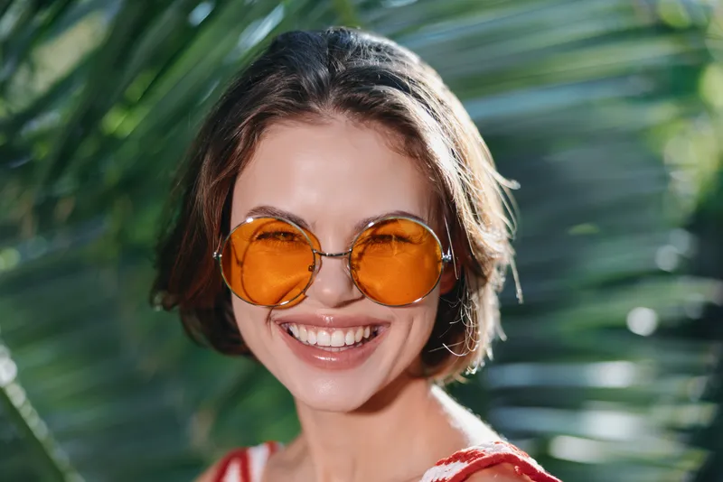 A cheerful portrait features a stylish woman wearing round orange sunglasses, smiling warmly at the camera with a sunny outdoor backdrop, evoking a vibrant, contemporary vibe.