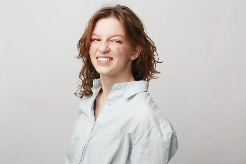Young woman with curly hair wearing a light shirt makes a playful grimace against a plain background. Concept of natural expression, personality, and casual lifestyle.