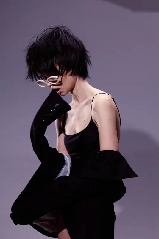 Stylish woman with short hair wearing black dress and white glasses in a modern fashion pose against gray background