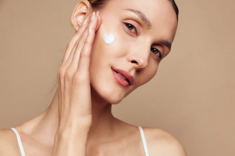 Young woman with clear skin applying white face cream on cheek. Close-up portrait with soft lighting on beige background. Concept of skincare, beauty, and daily routine.