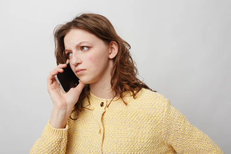 Young woman in yellow sweater talks on smartphone with serious expression, looking away. Isolated on light background. Concept of communication, concern, and thoughtful conversation.