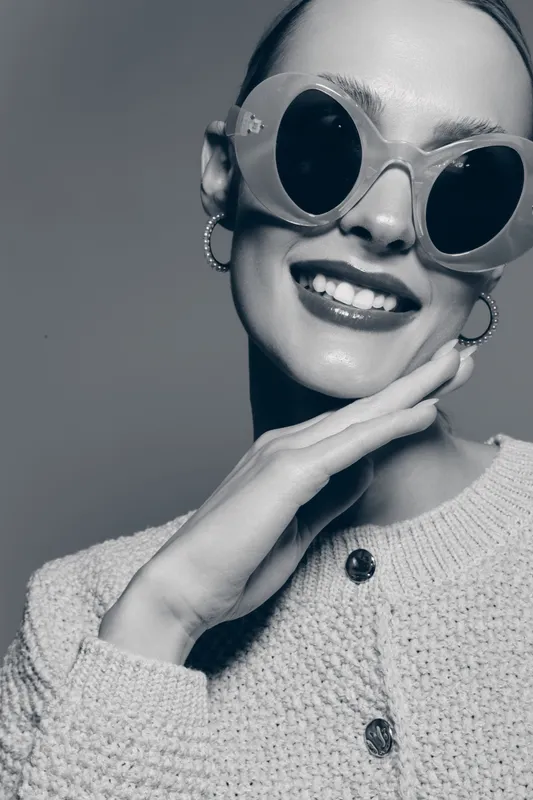 Smiling caucasian woman with pale skin wearing round sunglasses and hoop earrings poses with hand near face. Stylish portrait in black and white with cozy knitted sweater and confident mood.