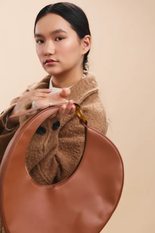 Asian woman wearing a warm brown coat holds a large leather bag and faces the camera with a calm, stylish expression, creating a refined fashion portrait in soft studio light.