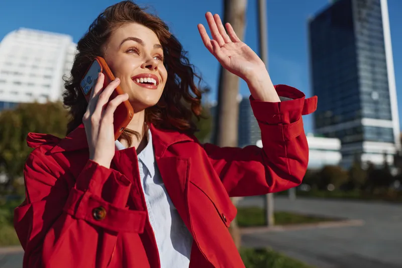 A young woman in a red coat talks on a smartphone outdoors, smiling with one hand raised as tall buildings rise behind her.