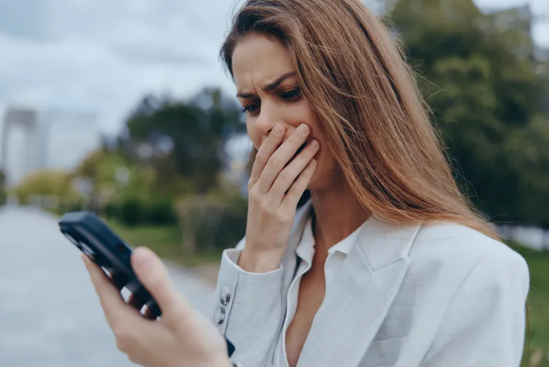Woman stressed by financial crisis holding phone outdoor worried about stock market fall in white blazer autumn