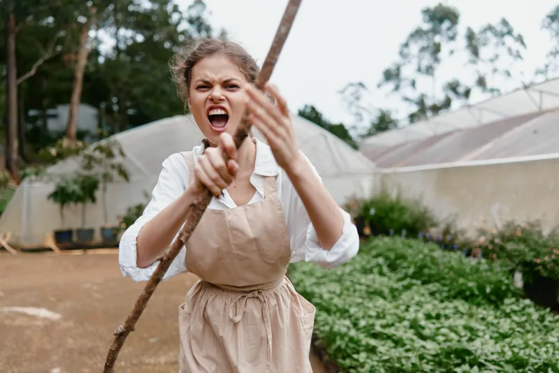 Young woman expressing anger while holding a stick in a garden, wearing a beige apron and white shirt, surrounded by greenery and greenhouses