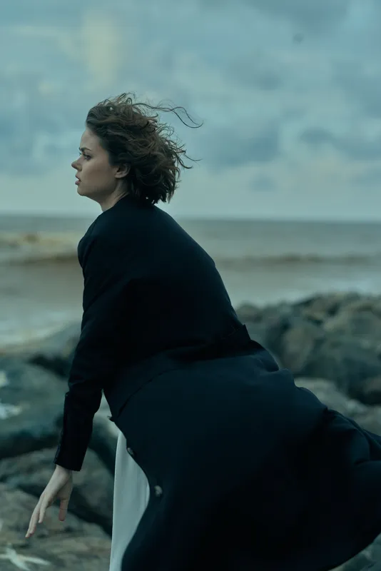 Moody scene of a woman on a windswept rocky shore, wearing a dark dress, facing the sea with flowing hair, a contemplative pose and dramatic atmosphere.