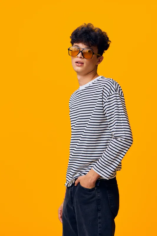 Young man wearing striped shirt and yellow sunglasses stands confidently against bright orange background, showcasing casual style and modern fashion trends.