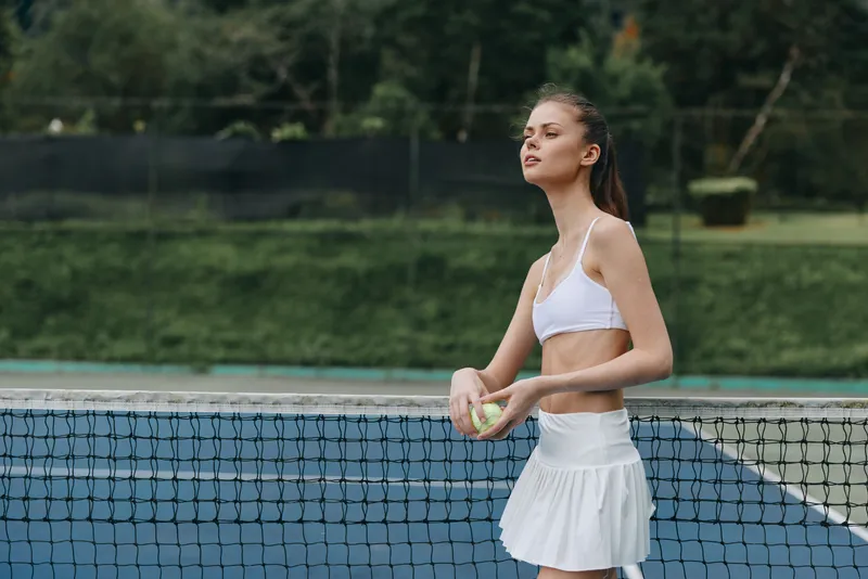 Young female athlete in a white sports outfit holding a tennis ball, exuding confidence and determination on a sunny outdoor tennis court