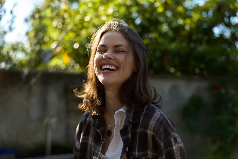 Smiling woman with shoulder length hair outdoors on a sunny day, wearing casual plaid shirt, natural light enhances happiness and relaxed lifestyle in a green garden