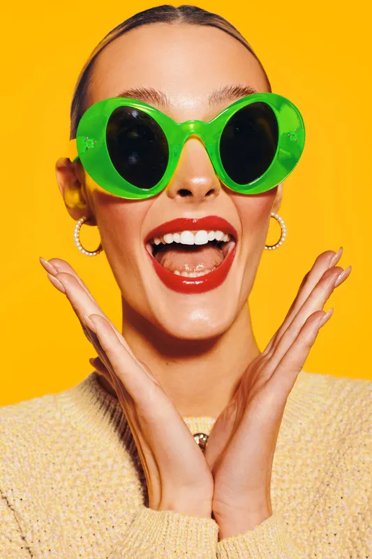 Happy young woman with bright red lipstick wearing large green sunglasses and beige sweater poses excitedly on yellow background. Concept of summer fashion, joy, and vibrant style.