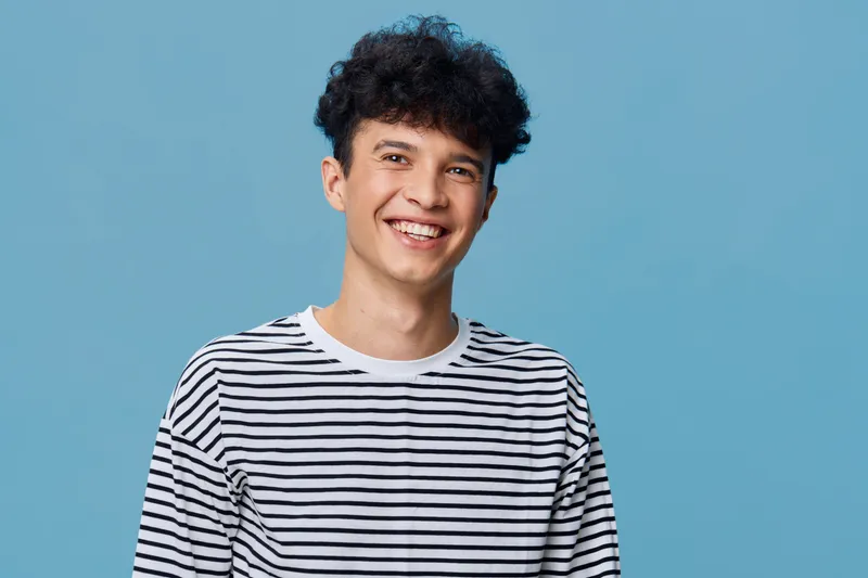 smiling man wearing striped shirt stands against solid blue background, joyful expression with curly hair, casual style portrait of young adult male