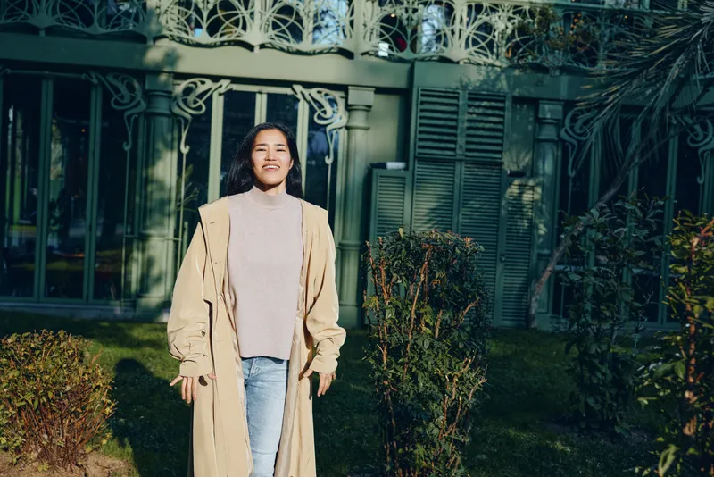 Woman in a beige coat and light turtleneck stands in a sunlit garden with a green fence, smiling, casual street style, candid outdoor fashion scene for lifestyle photography